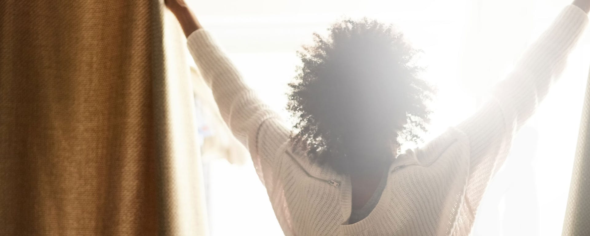 Une femme ouvrant ses rideaux pour laisser briller la lumière dans sa chambre.