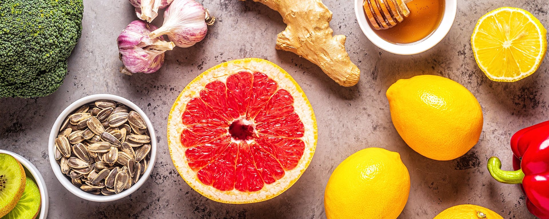 Top-down shot of a countertop with broccoli, lemons, grapefruit, ginger, red pepper, kiwi, sunflower seeds, and honey.