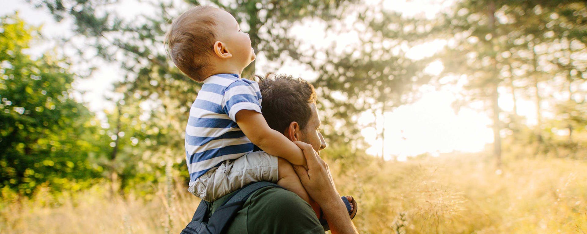 A father walking in the woods with his son on his shoulders.